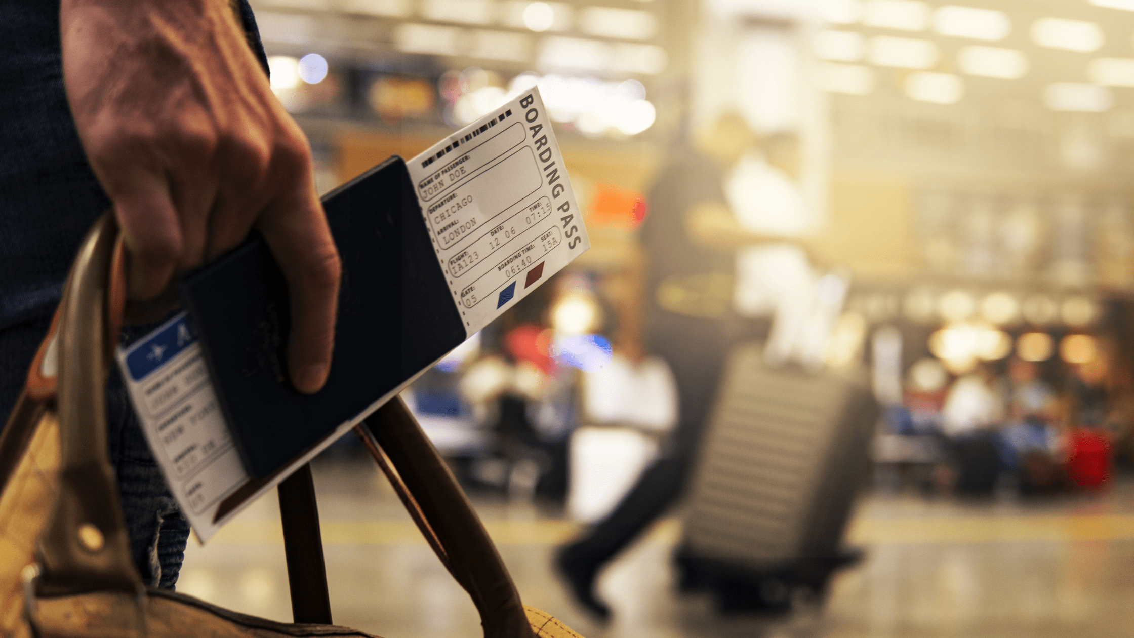 Shot of a traveler's hand holding a plane ticket tucket in a passport. Blurry background depicts a busy airport.
